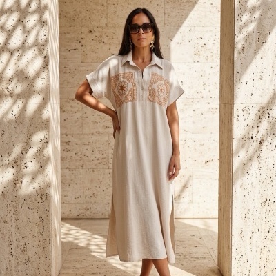 Mulher com vestido comprido bege e sandálias castanhas numa parede de pedra clara