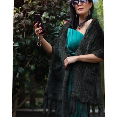 Mulher veste vestido verde-água e estola cinza escuro com franjas ao ar livre junto a plantas