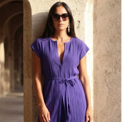 Mulher com vestido ou conjunto roxo canelado, óculos de sol escuros e brincos prateados, com parede de pedra clara ao fundo