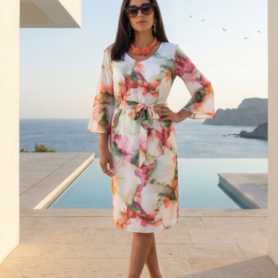 Vestido midi floral com cinto e mangas 3/4, colar coral e sandálias nude ao lado de piscina com vista para o mar