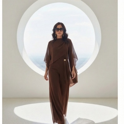 Macacão castanho com mangas transparentes e fivela dourada usado por mulher com óculos escuros frente a janela circular