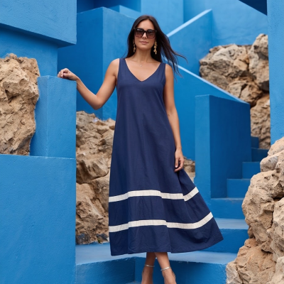 Mulher com vestido comprido azul escuro com duas faixas bege, sandálias transparentes, óculos escuros, em escadaria azul com pedras.