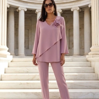 Mulher vestindo conjunto cor-de-rosa com blusa decorada e calças, em escadaria de mármore com colunas ao fundo.