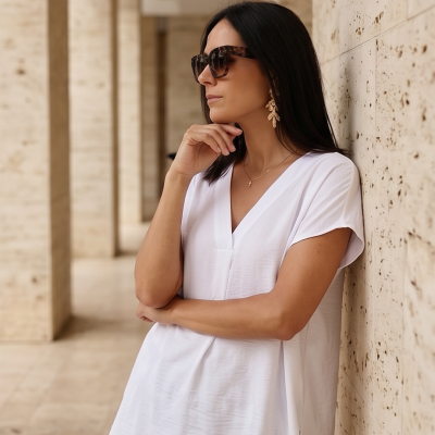 Mulher com óculos de sol, brinco dourado e blusa branca encostada a parede clara