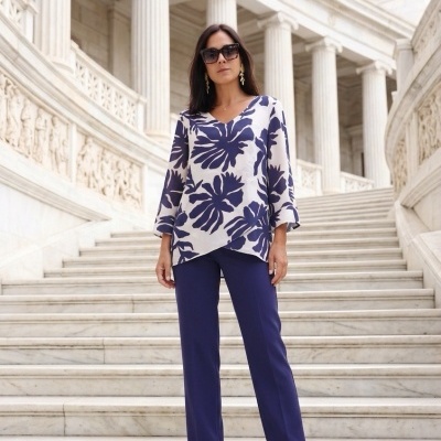 Mulher com blusa branca e azul com padrão de folhas, calças azuis e sapatos de salto em escadaria de mármore
