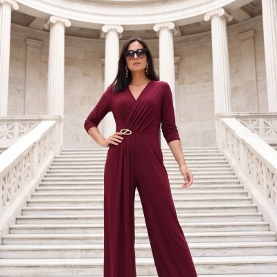 Mulher de macacão vinho com cinto dourado, óculos escuros e sandálias em escadaria com colunas brancas.
