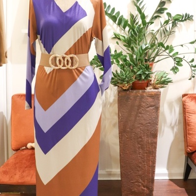 Vestido colorido comprido com cinto bege em manequim dentro de uma sala com plantas e cadeiras laranja.