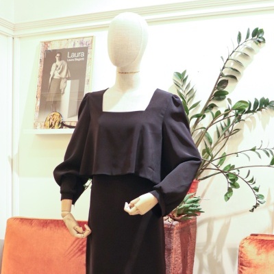 Vestido preto de mangas largas num manequim de tecido bege com sofá laranja e plantas ao fundo