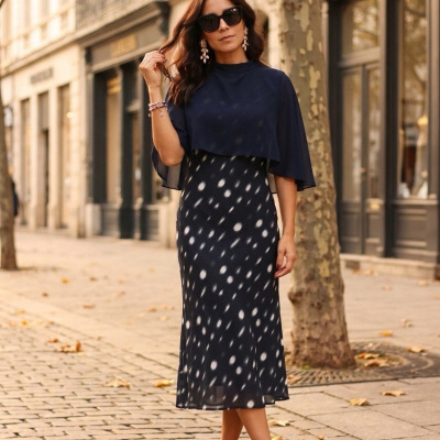 Mulher com vestido midi azul escuro de bolas brancas e acessórios numa rua com folhas caídas
