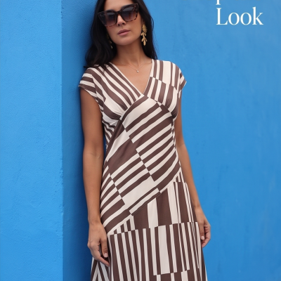 Mulher com vestido às riscas castanho e creme, óculos de sol e brincos dourados, encostada a parede azul com texto Shop the Look.
