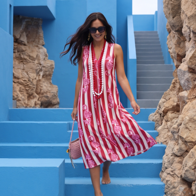 Mulher sorridente com vestido às riscas vermelhas, brancas e cor-de-rosa, óculos de sol e colares, entre escadas e paredes azuis