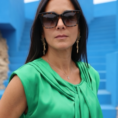 Mulher com óculos de sol castanhos, brincos dourados, colar prateado e blusa verde em escadas azuis.