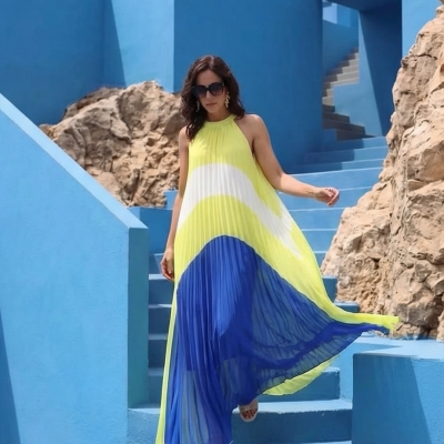 Vestido longo amarelo, branco e azul com tecido plissado, usado por mulher num cenário azul com escadas e pedras