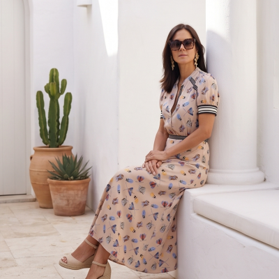 Mulher sentada com vestido comprido bege estampado e sandálias de cunha ao lado de coluna branca e vasos com plantas