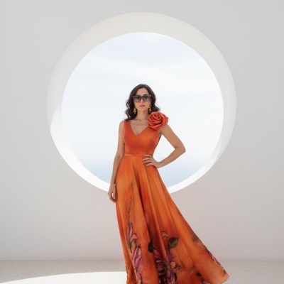 Vestido longo laranja com flores pintadas e detalhe de flor no ombro junto a modelo com óculos escuros em fundo branco com janela circular