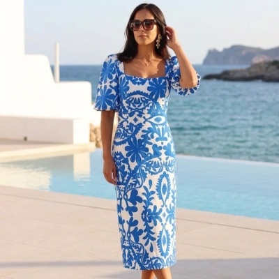 Mulher com vestido midi branco e azul com padrão floral junto a piscina com vista para o mar.