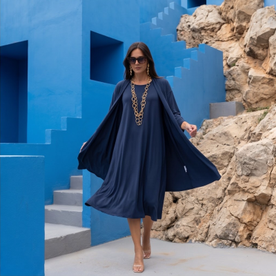 Mulher com vestido e casaco azul escuro e acessórios dourados junto a paredes azuis e pedras naturais