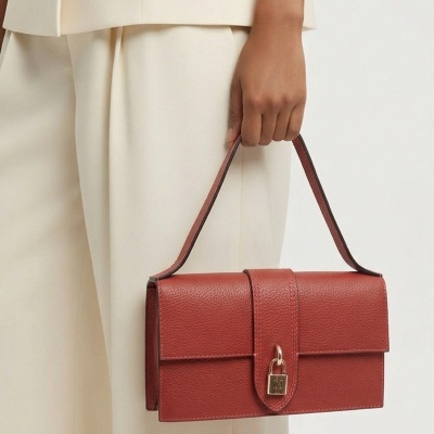 Mala de mão vermelha em pele com fecho de cadeado dourado, segurada por mão de pessoa vestindo roupa bege