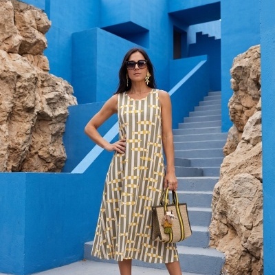 Mulher com vestido midi de padrão geométrico bege, amarelo e cinzento, sandálias de corda, bolsa estampada castanha e amarela, brincos dourados e óculos escuros, com fundo de paredes azuis e pedras.