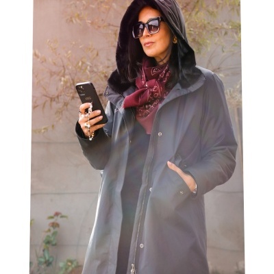 Mulher com casaco preto e capuz, lenço bordô e óculos de sol a usar telemóvel.