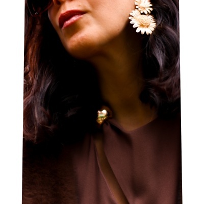 Mulher com brincos dourados em forma de flores e roupa castanha com botão dourado.