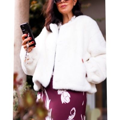 Mulher com casaco branco felpudo, saia bordô com padrão floral, óculos escuros e telemóvel preto com cordão