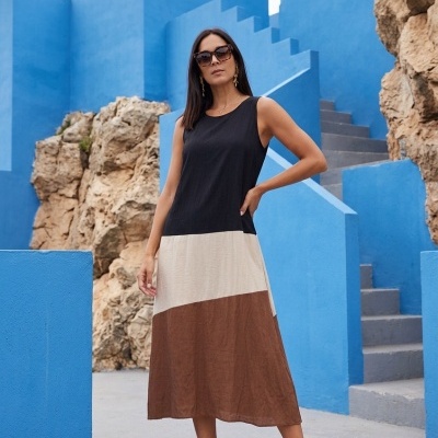 Mulher com vestido de blocos de cor preto, bege e castanho, sandálias douradas e óculos de sol em ambiente com paredes azuis.