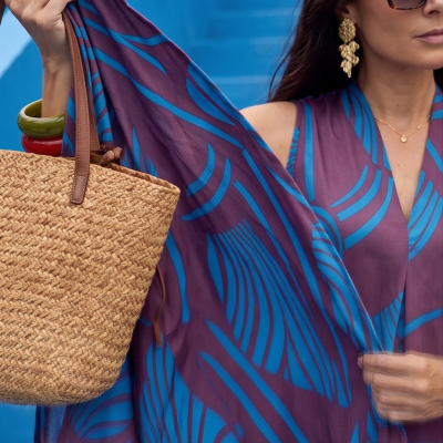 Mulher com vestido/xale azul e roxo segurando mala de palha com alças de couro castanho.