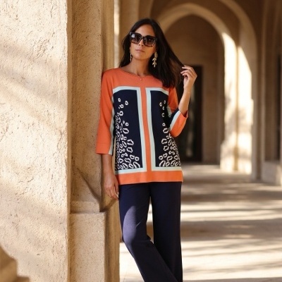 Mulher vestida com camisola colorida e calças azuis escuras junto a coluna em arco de pedra