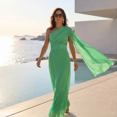 Mulher com vestido verde longo de um ombro e capa a caminhar junto a piscina com vista para o mar