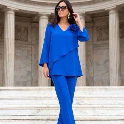 Mulher de azul royal com blusa e calças, sandálias nude, óculos de sol e brincos dourados em escadaria de mármore.