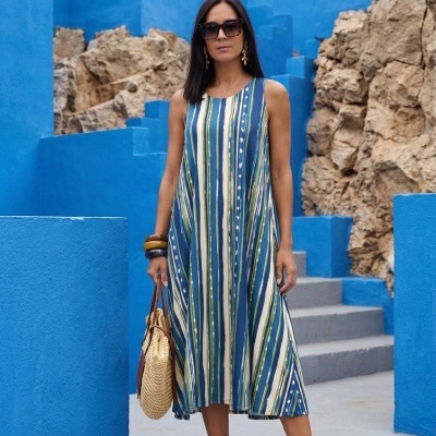 Mulher com vestido de riscas verticais azul, verde e branco, sandálias beges, bolsa de palha, em ambiente azul com pedras e escadas.