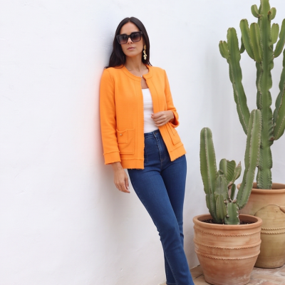 Mulher com casaco laranja, jeans azul e óculos escuros, ao lado de cactos em vasos de barro.