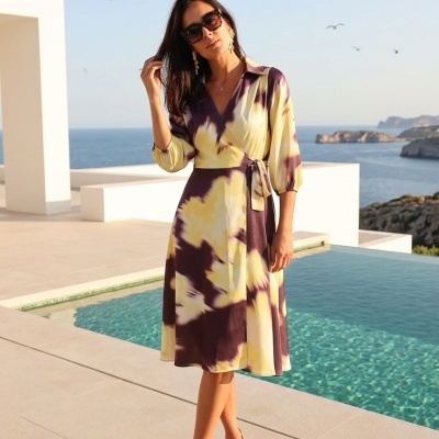 Mulher com vestido midi tie-dye amarelo e roxo junto à piscina com mar ao fundo