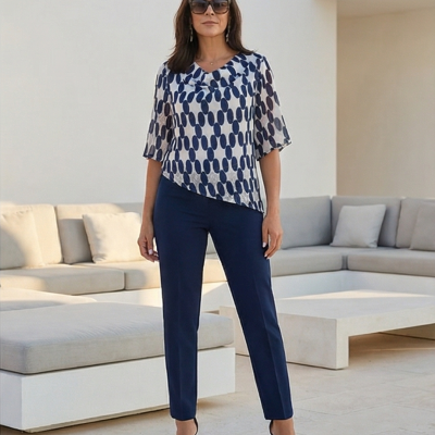 Mulher vestindo blusa branca com manchas azuis, calças azul marinho, sandálias de salto, óculos escuros, em espaço exterior com móveis claros.