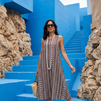 Mulher a usar vestido midi castanho com padrão branco, sandálias castanhas, óculos de sol e mala de palha em escadaria azul.