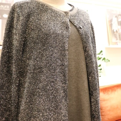 Cardigan cinza escuro felpudo com botões em manequim, vestido cinza claro por baixo, interior com sofá laranja