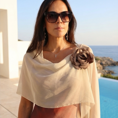 Mulher com capa bege transparente e flor decorativa castanha no ombro perto de piscina com vista para o mar