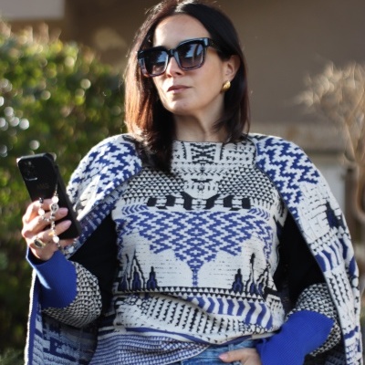 Mulher com camisola de malha azul, branco e preto, casaco e jeans azuis, óculos de sol pretos grandes, brincos dourados e telemóvel.