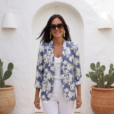Blazer azul com flores brancas, usado por mulher com óculos e brincos dourados, parede branca e vasos com cactos ao fundo