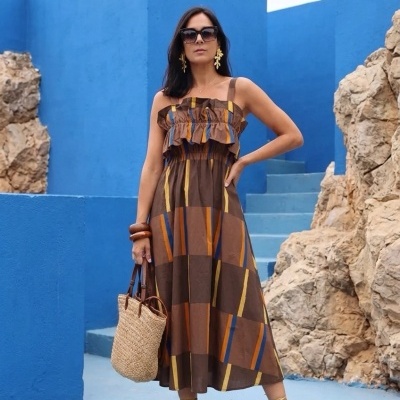 Mulher com vestido midi colorido e mala de palha diante de parede azul
