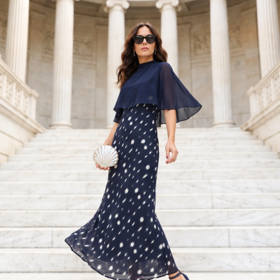 Mulher com vestido azul escuro de manchas brancas e capa transparente, sandálias e mala formato concha, em escadaria de mármore branco com colunas altas