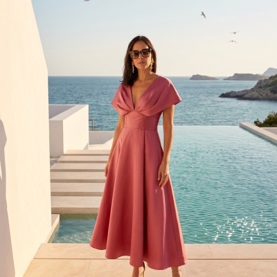 Mulher com vestido rosa midi e óculos escuros junto a piscina com vista para o mar.