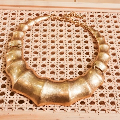 Colar dourado segmentado colocado sobre superfície de padrão entrelaçado em madeira clara