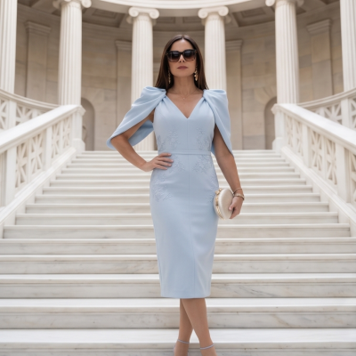 Mulher de vestido midi azul claro posando numa escadaria de mármore branco com colunas clássicas, usando óculos de sol pretos e segurando bolsa bege.