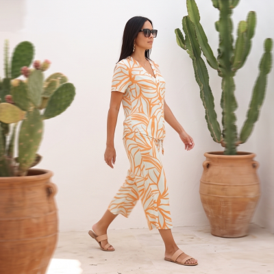 Conjunto de roupa feminina bege e laranja com padrão de folhas usado por mulher ao lado de vasos de barro com cactos