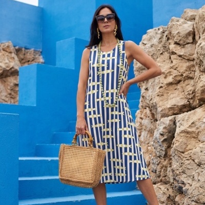 Mulher com vestido midi azul, branco e amarelo, sandálias castanhas e mala de palha em escadaria azul