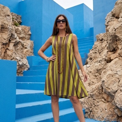 Mulher com vestido às riscas verdes e castanhas, óculos escuros e sandálias douradas em escadaria azul com pedras ao lado.