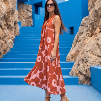 Mulher com vestido floral laranja e rosa, sandálias castanho, colar de contas laranja e óculos de sol, em escadaria azul entre rochas