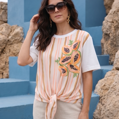 Mulher com t-shirt branca com riscas e estampa de papaias, calças claras, escadas azuis e rochas ao fundo.